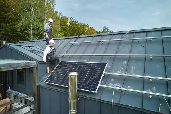 Men workers prepearing for mounting photovoltaic solar modules on roof of house. Electricians in helmets installing solar panel system outdoors. Concept of alternative and renewable energy.