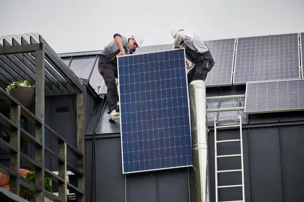 Men roofers installing solar panel system on roof of house. Technicians in helmets lifting up photovoltaic solar module with help of ropes outdoors. Concept of alternative and renewable energy.