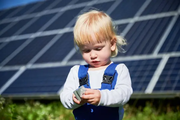 Little child looking at small detail of big solar station. Interested boy learning how to use tools. Cute blond kid holding small piece on background of solar panels at sunny day.
