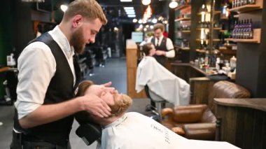 Profesyonel berber müşterilerin sakallarını tıraş ediyor. Süslü iç mimarisi olan modern bir berber dükkanı. İş yerinde zarif kıyafetli bir kuaför..