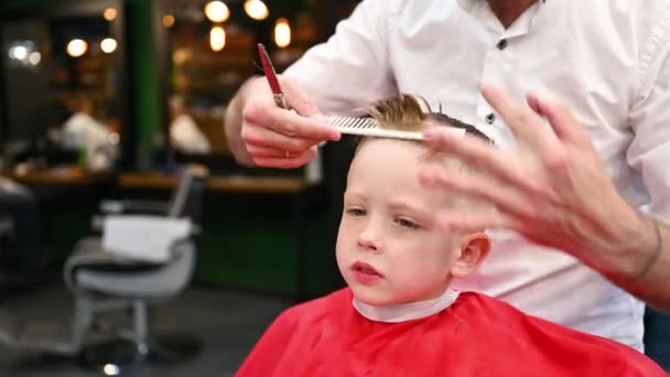 男人理发师用梳子和剪刀剪小男孩的头发 接近孩子从成年男性那里理发 很可能是理发师 现代理发店的专业理发师和可爱的顾客 — 图库视频影像