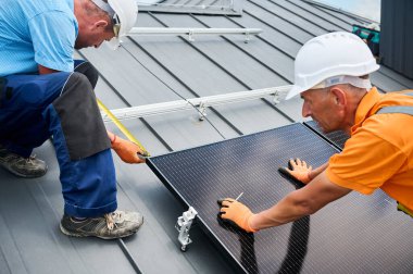 İşçiler fotovoltaik etkilerle elektrik üretmek için evin çatısına güneş paneli inşa ediyorlar. Kurulum için montaj ekipmanlarını ölçmek için cetvel kullanan adamlar.