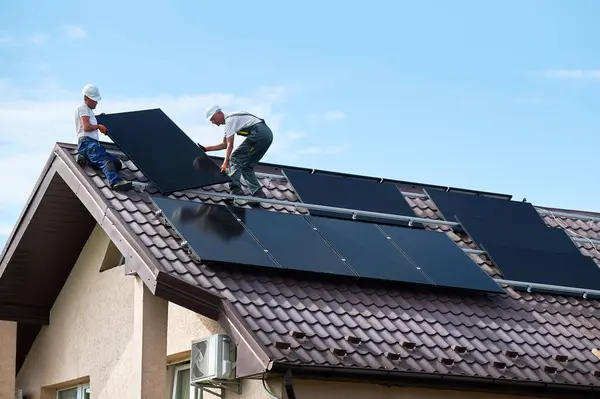 Workers building solar panel system on roof of house. Two men installers in helmets carrying photovoltaic solar module outdoors. Alternative, green and renewable energy generation concept.
