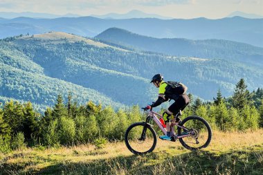Dışarıda elektrikli dağ bisikleti süren bisikletçi. Erkek turist dağlarda otlak patika boyunca bisiklet sürüyor, başlık ve sırt çantası takıyor. Spor anlayışı, aktif eğlence ve doğa.