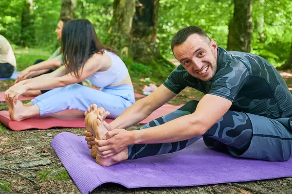 People engaged in stretching exercise outdoors, part of yoga practice. Portrait of happy man stretching forward to reach his toes, sitting on purple yoga mat.