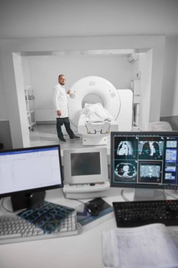 Tıbbi bilgisayarlı tomografi ya da MRI tarayıcısı. Doktor MR çekiyor, hasta kanepede yatıyor. Beyaz cüppeli ve gözlüklü bir erkek uzman kameraya bakıyor, gülümsüyor. Modern teşhis kavramı.