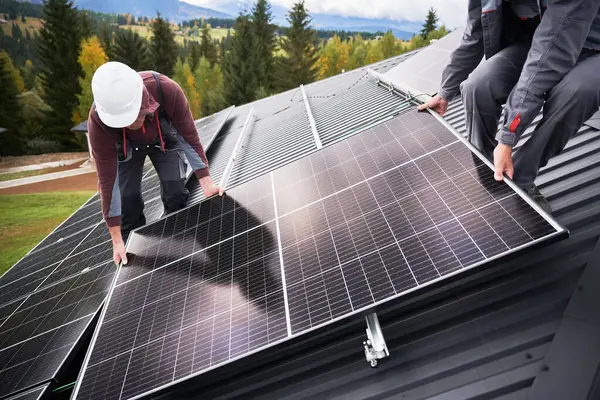 Builders building photovoltaic solar module station on roof of house. Men electricians in helmets installing solar panel system outdoors. Concept of alternative and renewable energy.