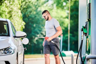 Benzin pompalı bir sürücü benzin deposunu dolduruyor. Lüks beyaz otomobiline yakıt dolduran kendinden emin bir adam. Modern benzin istasyonunda günlük kıyafetler ve güneş gözlüğü takan bir adam..