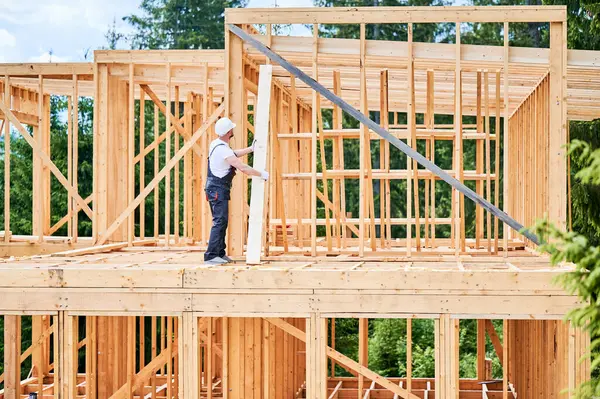 Carpenter constructs wooden-framed house. Man holds large plank in his hands while dressed in work clothes and helmet. Concept of modern ecological construction.