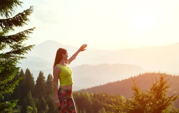 Portrait of sporty, young female traveling, hiking in mountains. Slim woman wearing sportswear, standing on hill, raising hand, smiling, enjoying. Concept of harmony with nature.