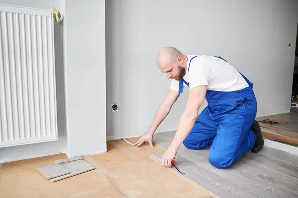 Man in work overalls measuring distance from wall to laminate board. Male construction worker using tape measure while installing laminate flooring in apartment under renovation.
