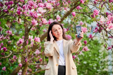 Mutlu kadın akıllı telefonuyla selfie çekiyor bahar çiçekleri arasında arka planda yumuşak bir odak ile.