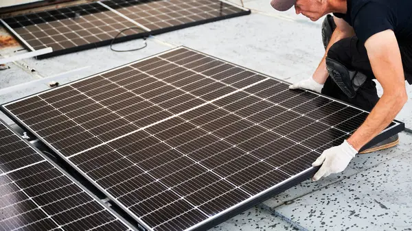 Worker building solar panel system on metal rooftop of house. Man engineer in gloves installing photovoltaic solar module outdoors. Alternative, green and renewable energy generation concept.