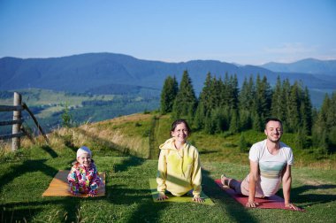 Genç bir aile sabahları doğa manzaralı dağlarda yoga yapıyor. Baba, anne ve kız yoga minderlerinde, arka planda dağları ve ormanı manzaralı açık mavi gökyüzünün altında.