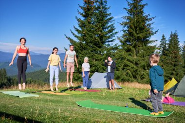 Yoga yapan bir grup insan dağlarda kamp yaparken dışarıda poz veriyor. Yetişkinler ve çocuklar yoga minderlerinin üzerinde duruyor, her biri sabah açık mavi bir gökyüzünün altında yoga pozu veriyor. Genç çocuk eğitmendir..