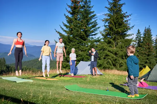 Group of people doing yoga pose outdoor in camping in the mountains. Adults and children standing on yoga mats, each doing a yoga pose under a clear blue sky in the morning. Young boy is instructor.
