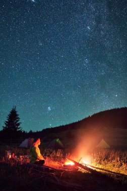 Yıldızlı gökyüzünün altındaki dağlarda gece kampı. Kamp ateşinin yanında çimenlerde oturan bir kadın manzaraya hayran kalıyor, dinleniyor, eğleniyor. Doğayla uyum kavramı.