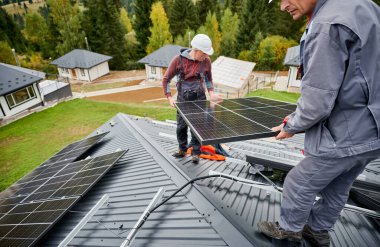 Evin çatısına güneş paneli inşa eden mühendisler. Dışarıda fotovoltaik güneş modülü taşıyan miğferli işçiler. Alternatif ve yenilenebilir enerji kavramı.