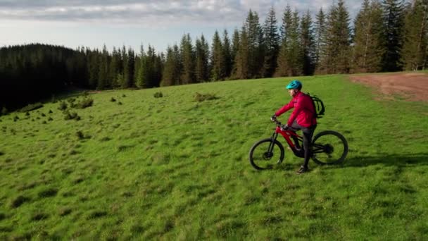 サイクリストの男性は 晴れた夜に屋外で電動自転車に乗っています 美しい山の風景を楽しむ草の丘に休む男性の観光客は ヘルメットとバックパックを着ています アクティブレジャーのコンセプト — ストック動画