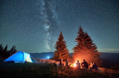 Night camping in mountains under starry sky. Group of people tourists having a rest near campsite, burning campfire and illuminated tent. Concept of tourism, hiking and adventure.
