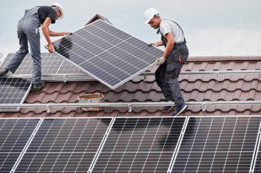 Erkek teknisyenler evin çatısında fotovoltaik güneş modülü taşıyorlar. Kasklı elektrikçiler dışarıda güneş paneli sistemi kuruyorlar. Alternatif ve yenilenebilir enerji kavramı.