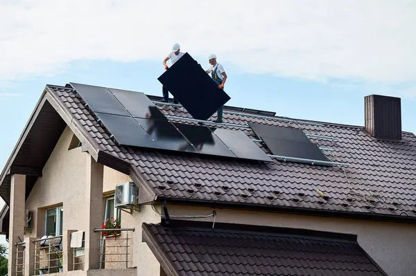 Workers building solar panel system on roof of house. Two men installers in helmets carrying photovoltaic solar module outdoors. Alternative, green and renewable energy generation concept.