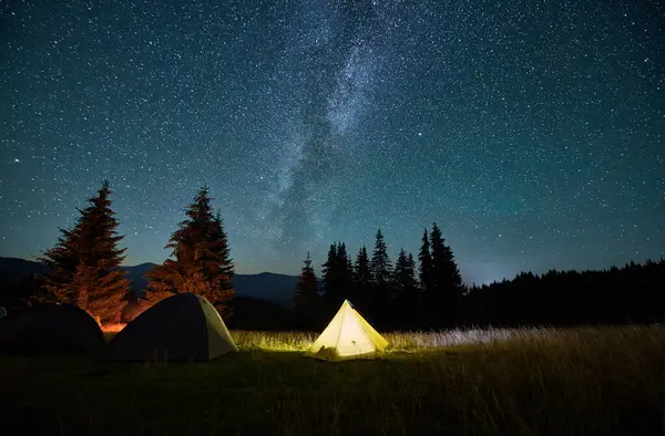 Night camping in mountains under starry sky. Tourist tents in campsite near burning campfire under beautiful sky full of stars with Milky way above forest. Concept of tourism and traveling.