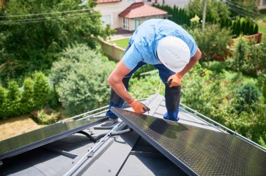 Evin çatısında fotovoltaik güneş paneli inşa eden bir işçi. Miğferlerde mühendis ve eldivenler dışarıda altıgen anahtarla güneş modülü kuruyor. Yenilenebilir enerji üretimi kavramı.