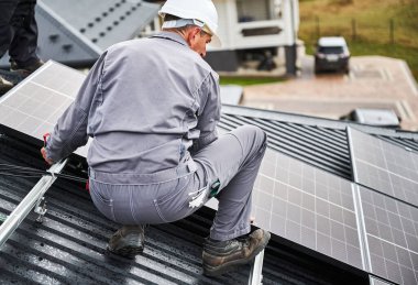 Evin çatısına fotovoltaik güneş modülleri monte eden bir inşaatçı. Güneş paneli sistemi kuran miğferdeki insan mühendisin arka görüntüsü. Alternatif ve yenilenebilir enerji kavramı.
