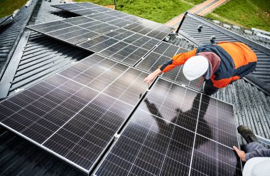 Man builder mounting photovoltaic solar panels on roof of house. Engineer in helmet installing solar module system with help of hex key. Concept of alternative, renewable energy. clipart