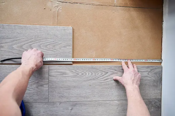 Close up of male construction worker using tape measure ruler while measuring distance from wall to laminate board. Man installing laminate flooring in apartment under renovation.