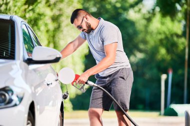 Benzin pompalı bir sürücü benzin deposunu dolduruyor. Lüks beyaz otomobiline yakıt dolduran kendinden emin bir adam. Modern benzin istasyonunda günlük kıyafetler ve güneş gözlüğü takan bir adam..