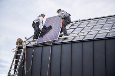 Elektrikçiler evin çatısına güneş paneli kuruyorlar. Dışarıda fotovoltaik güneş modülü taşıyan miğferli işçiler. Alternatif ve yenilenebilir enerji kavramı.