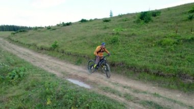 Dışarıda elektrikli dağ bisikleti süren bisikletçi. Erkek turistin dağlardaki otlak patika boyunca bisiklet sürüşünü, kaskını ve sırt çantasını gösteren hava manzarası. Spor anlayışı, aktif eğlence ve doğa.