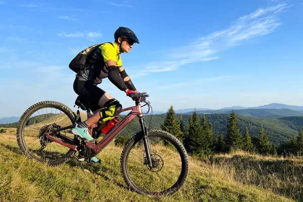 stock image Cyclist man riding electric mountain bike outdoors. Male tourist biking along grassy trail in the mountains, wearing helmet and backpack. Concept of sport, active leisure and nature.