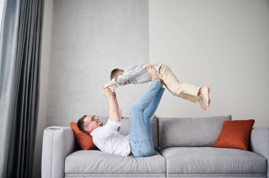Joyful father lifting happy little son at home. Carefree father and son weekend. Happy young father lying on couch and fooling around with kid.