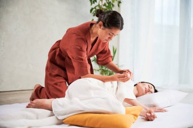 Masseuse making thai yoga massage. Woman therapist doing traditional massage treatment for female patient using elbow. Client lying on mat, meditating, enjoying hand massage in calm atmosphere.