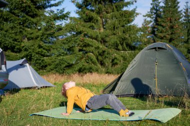 Kamptaki çim tarlasında yoga yapan genç çocuk yeşil minderde poz veriyor. Arka planda yeşil ağaçların arasına iki turist çadırı kuruldu..