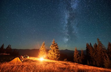 Yıldızlı gökyüzünün altındaki dağlarda gece kampı. Samanyolu ile dolu güzel gökyüzünün altındaki kamp alanında turist çadırları. Yürüyüşçülerin silueti yanan kamp ateşinin yanında dinleniyor. Turizm kavramı.