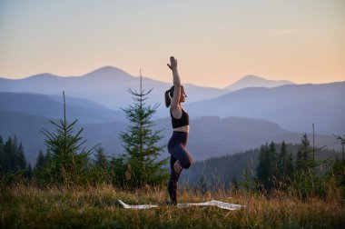 Dağlarda yoga yapan bir kadın, sakin, doğal bir ortamda. Kadın yoga pozu veriyor paspasın üzerinde, gün doğumunda ya da günbatımında güzel dağ manzarasının arka planıyla.