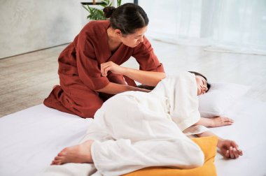 Masseuse making thai yoga massage. Woman therapist doing traditional massage treatment for female patient using elbow. Client lying on mat, meditating, enjoying hand massage in calm atmosphere.