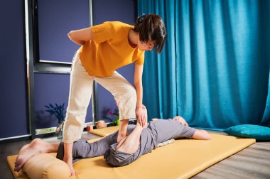 Masör Tayland yogası masajı yapıyor. Geleneksel masaj tedavisi uygulayan kadın terapist, erkek hastaların vücudunu esnetiyor. Müşteri paspasın üzerinde uzanıyor, sakin atmosferde el masajının keyfini çıkarıyor..