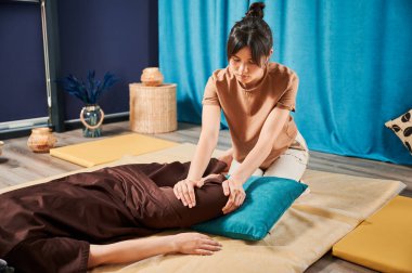 Masör Tayland yogası masajı yapıyor. Geleneksel masaj tedavisi uygulayan kadın terapist, kadın hastaların vücudunu geriyor. Müşteri paspasın üzerinde uzanıyor, sakin atmosferde el masajının keyfini çıkarıyor..