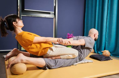 Masör Tayland yogası masajı yapıyor. Geleneksel masaj tedavisi uygulayan kadın terapist, erkek hastaların vücudunu esnetiyor. Kel müşteri paspasın üzerinde uzanıyor, sakin atmosferde el masajının keyfini çıkarıyor..