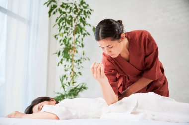 Masseuse making thai yoga massage. Woman therapist doing traditional massage treatment for female patient using elbow. Client lying on mat, meditating, enjoying hand massage in calm atmosphere.