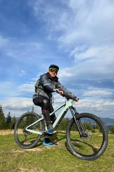 stock image Cyclist man riding electric bike outdoors on sunny day. Portrait of male tourist resting on grassy hill, enjoying beautiful mountain landscape, wearing helmet and backpack. Concept of active leisure.