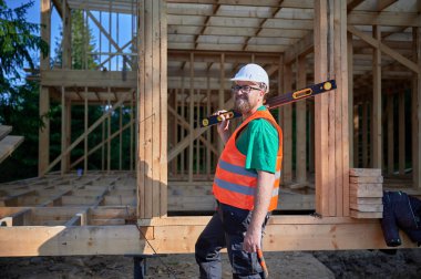 Marangoz ahşap çerçeve iki katlı ev inşa ediyor. Sakallı, gözlüklü, gözlüklü koruyucu kask takan, tulumlu ve turuncu yelek giyen bir adam. Modern ekolojik yapı kavramı.