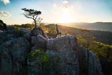 Yoga ve meditasyon yapan bir kadın akşamları dev bir kayanın tepesinde. Güzel günbatımı, sonbahar ormanları, arka planda kayalar ve tepeler. Hava aracı görünümü