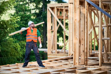 Marangoz ormanın yakınında ahşap çerçeveli ev inşa ediyor. Gözlüklü sakallı bir adam omuzunda büyük bir kiriş tutuyor, iş kıyafetleri ve miğfer giyiyor. Modern ve çevre dostu bina kavramı.
