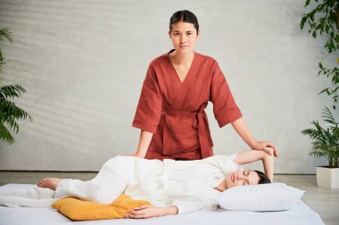 Masör Tayland yogası masajı yapıyor. Geleneksel masaj tedavisi yapan kadın terapistin portresi, kadın hastaların vücudunu germek. Müşteri paspasın üzerinde uzanıyor, sakin atmosferde el masajının keyfini çıkarıyor..
